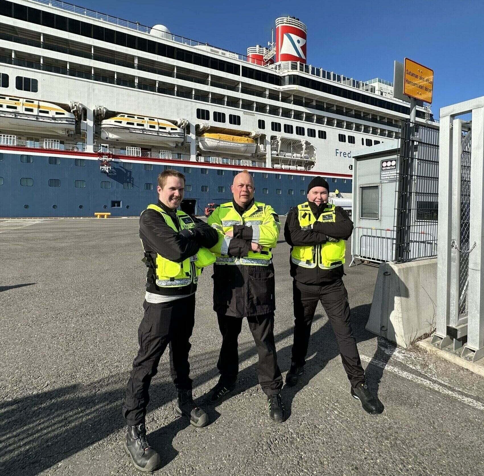 Kuvassa on kolme turvallisuusalan henkilöä. Taustalla näkyy risteilijä.