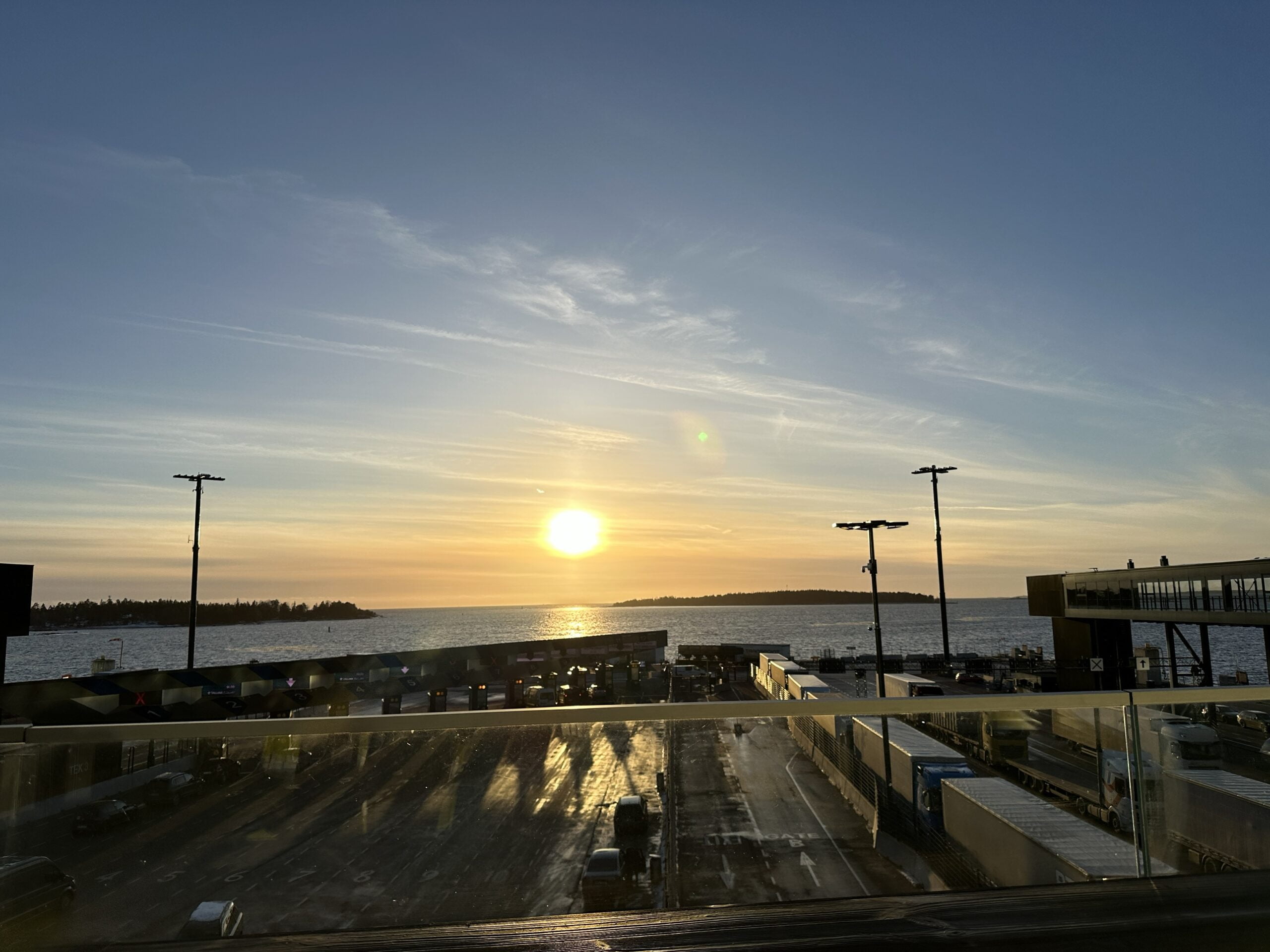 Kuvassa näkyy aurinko ja merta. Ollaan satamassa. Kuvassa on rekkoja.